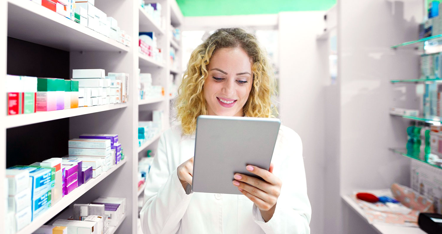A smiling pharmacist in a white coat uses a digital tablet while standing between shelves stocked with medications and health products in a bright, modern pharmacy.