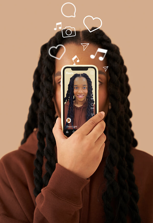 Person holding a smartphone in front of their face showing a selfie on a social media app, with illustrated icons like hearts and music notes floating above.